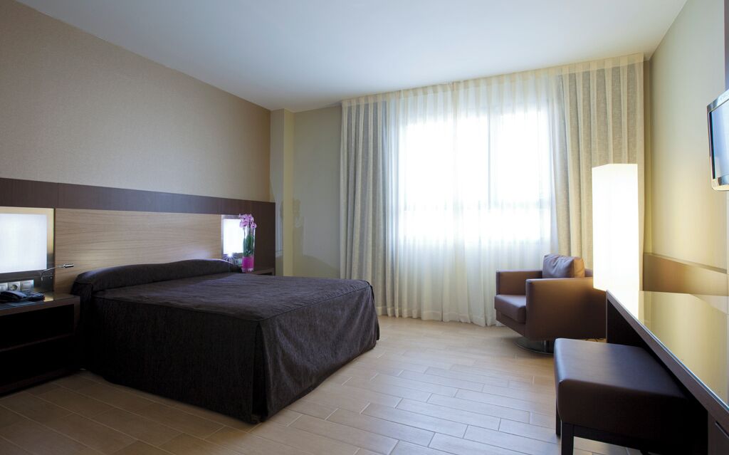 A hotel room with a bed, a desk, a chair, a TV, and a window with curtains.