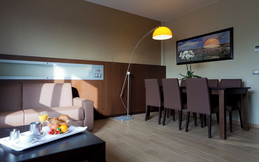 Una camera d'albergo moderna con divano, tavolo da pranzo, sedie e vassoio per la colazione con croissant, frutta e succo di frutta.