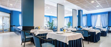 Un comedor con mesas dispuestas para el servicio, cortinas azules y un diseño de techo moderno.