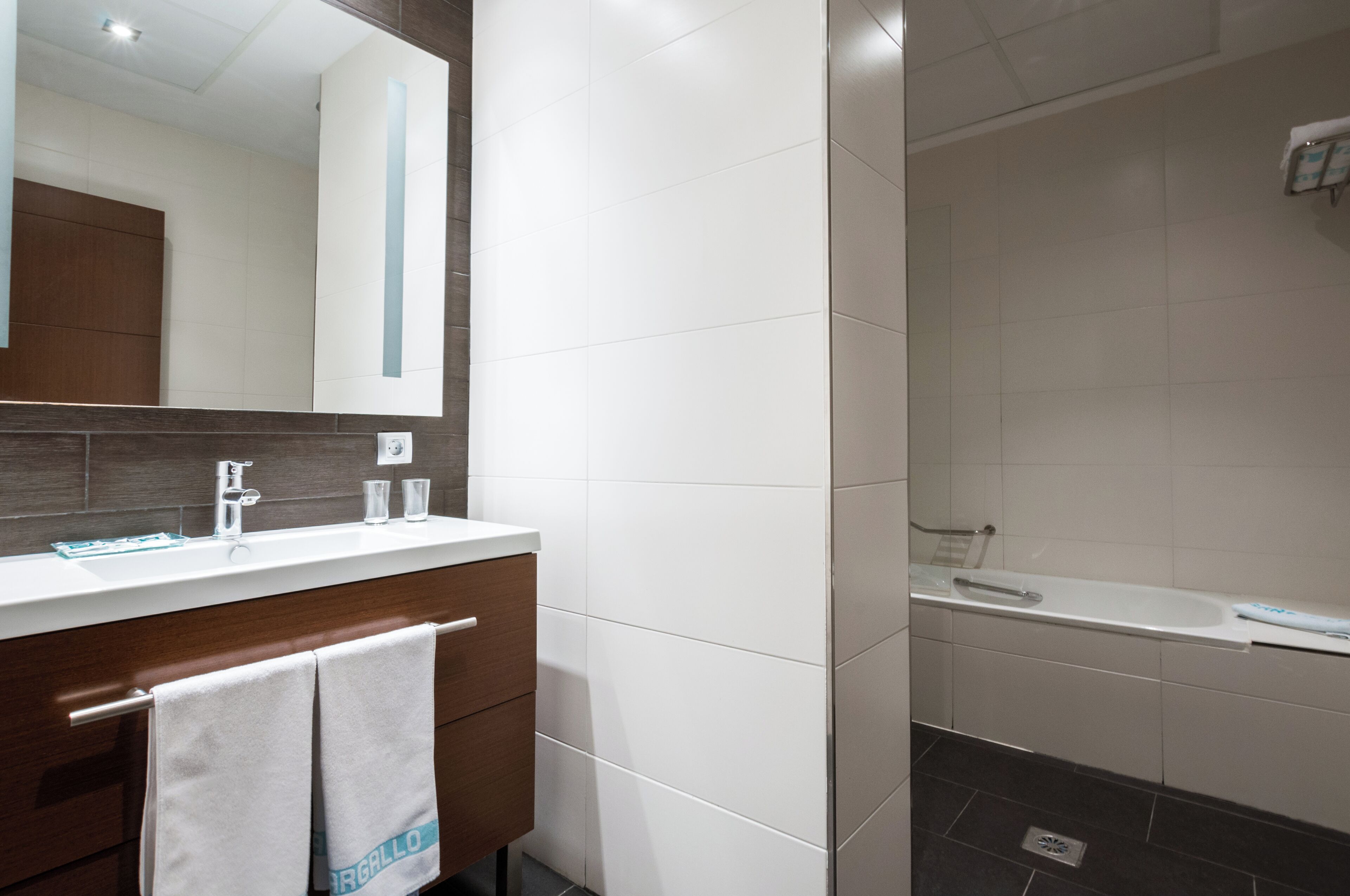 A modern bathroom with a white sink, mirror, and bathtub. There are towels hanging on the wall and a wooden vanity.