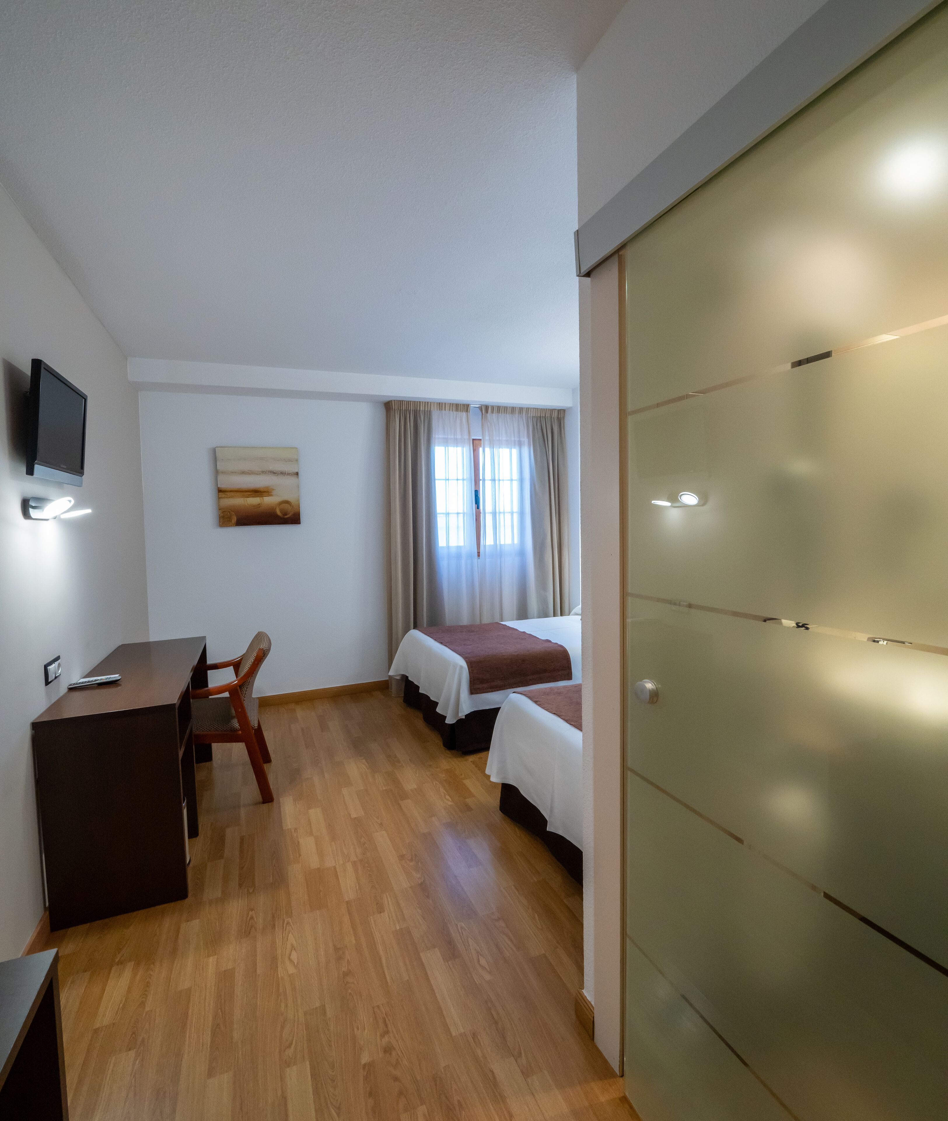A room with a wooden desk, two beds, a TV, and a painting on the wall.