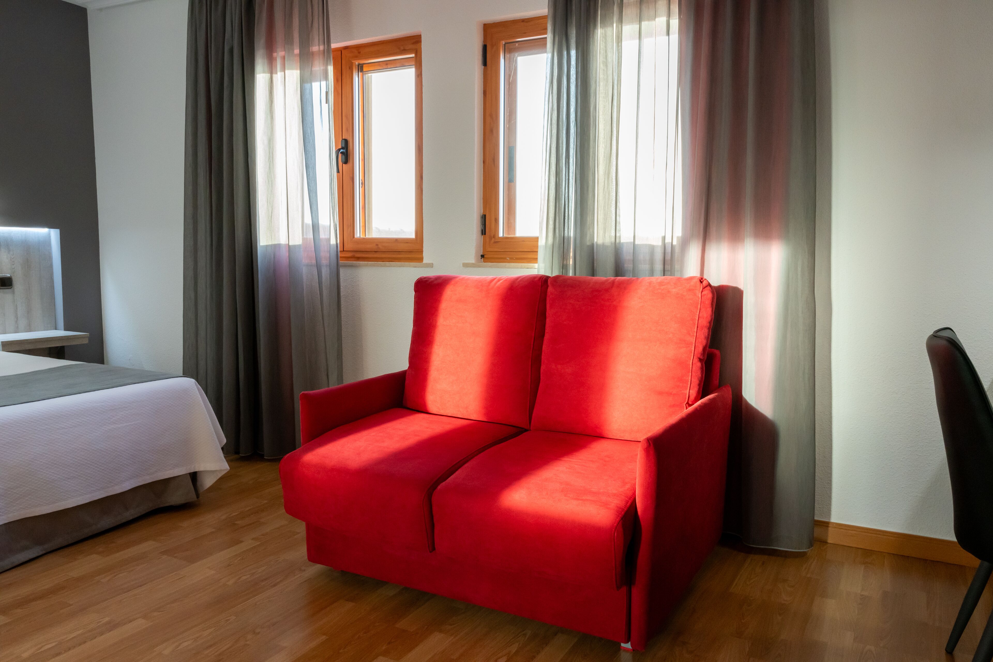 A modern room with a red sofa, a bed with a white duvet, and two windows with curtains.