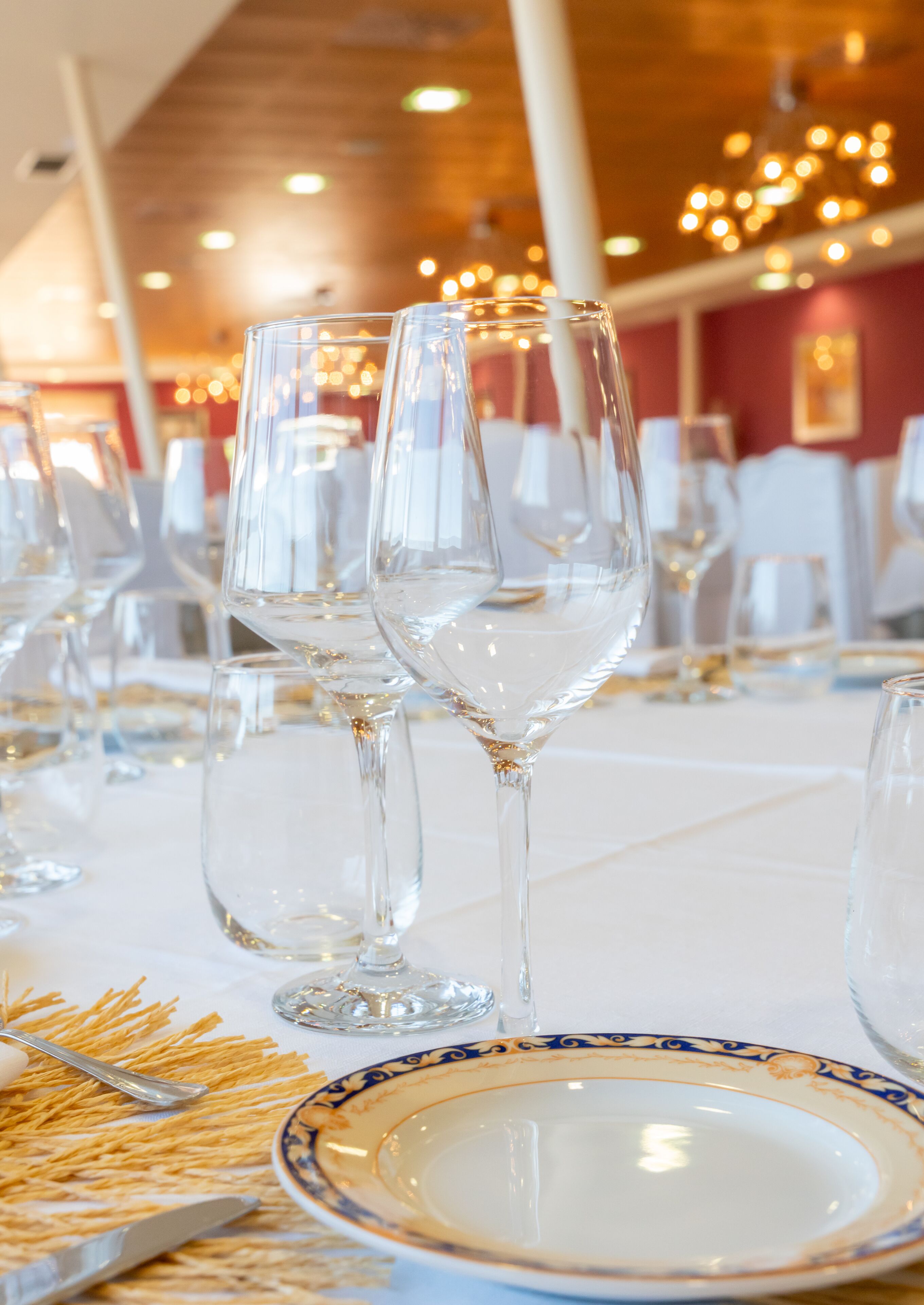 Une table à manger dressée avec des verres à vin transparents, une assiette à bordure bleue et dorée, et une cuillère posée sur un set de table en fibres tressées.
