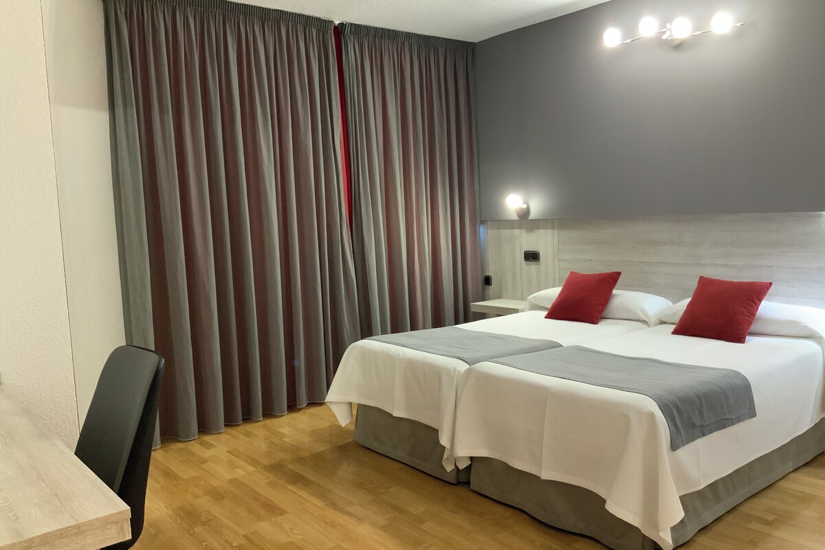 A hotel room with a double bed, two bedside tables, a desk with a chair, and a large window with curtains.