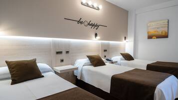 A hotel room with three beds, each with a brown headboard, white bedding, and a brown throw pillow. There is a wall-mounted light fixture above the beds and a wall art piece to the right.