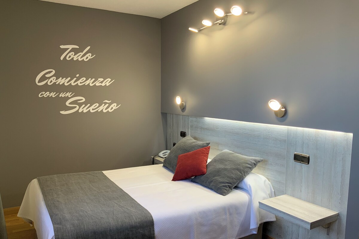 Une chambre moderne aux tons de gris et de blanc, avec un lit agrémenté d’oreillers gris et rouges, une table de chevet et un luminaire fixé au mur.