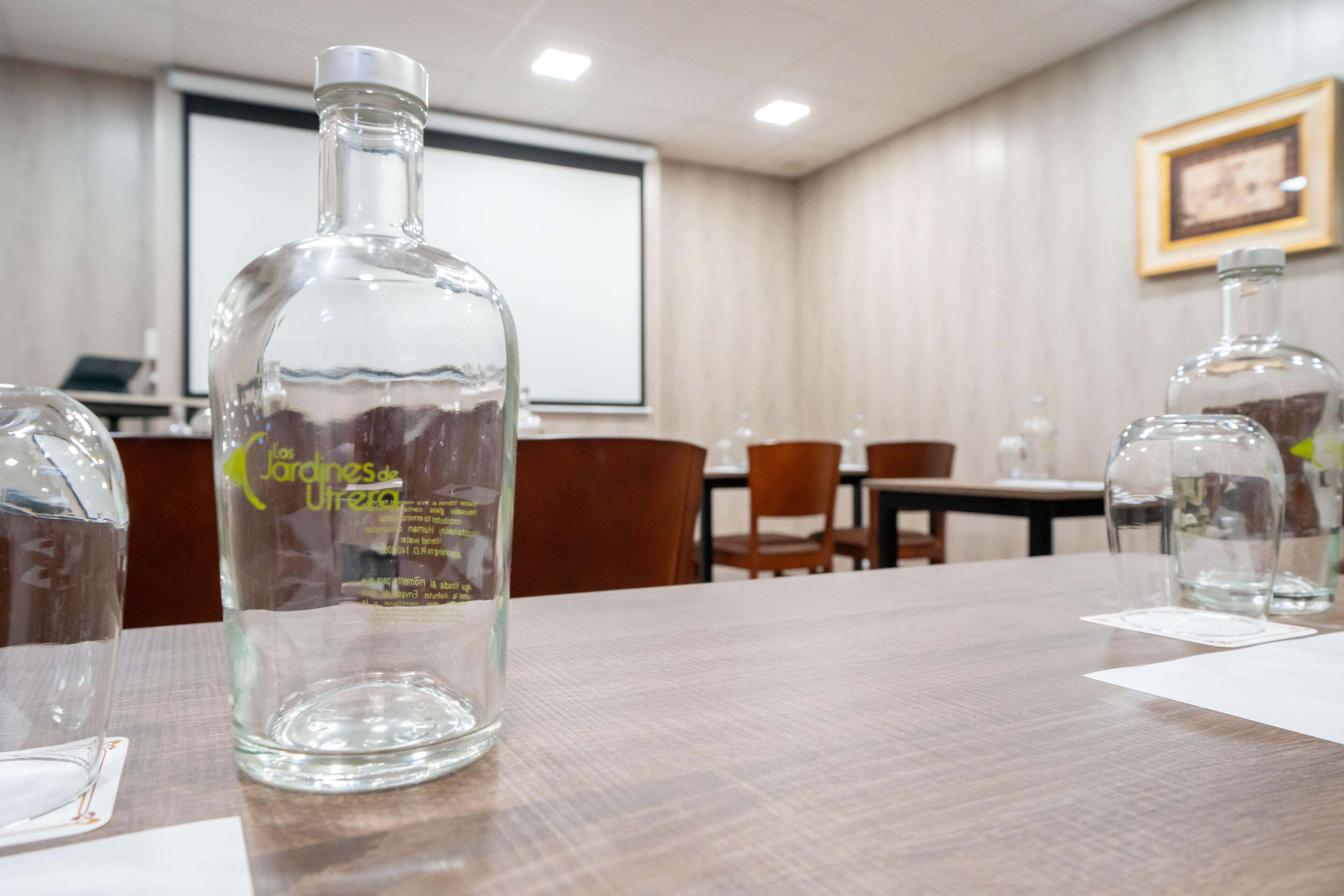 A conference room with a wooden table, clear glass bottles, and a projector screen in the background.