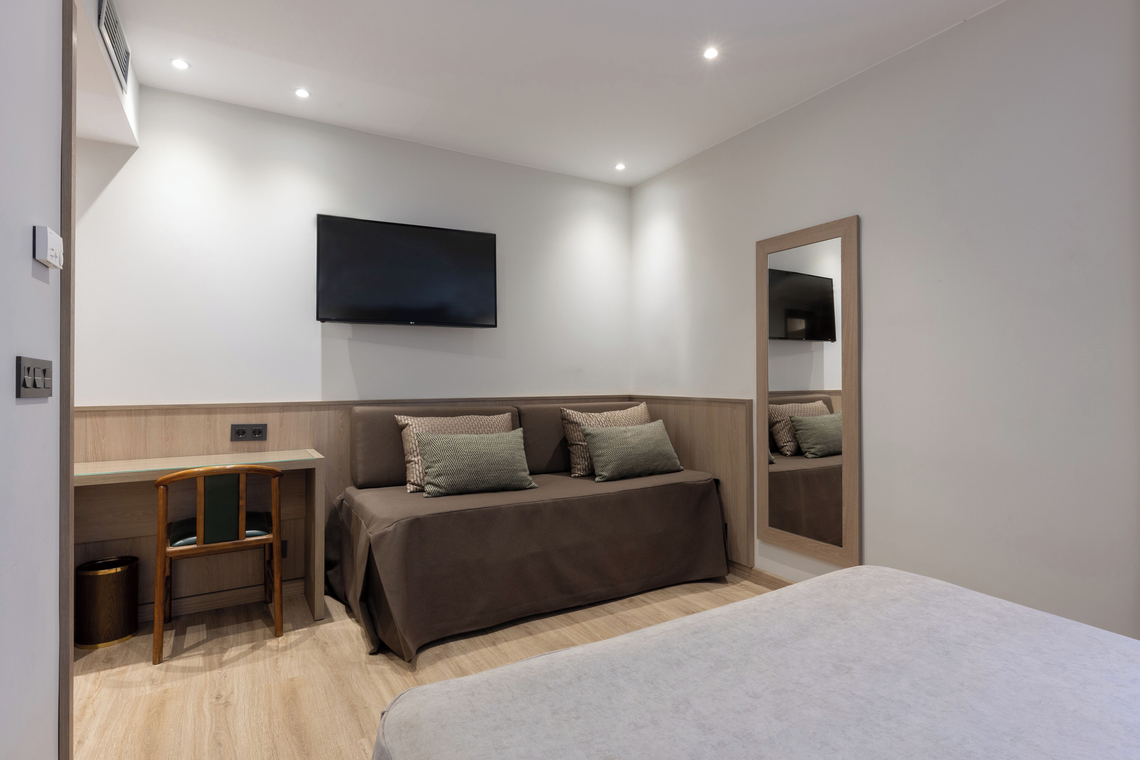 A modern hotel room with a bed, a desk, a chair, a television mounted on the wall, and a mirror.