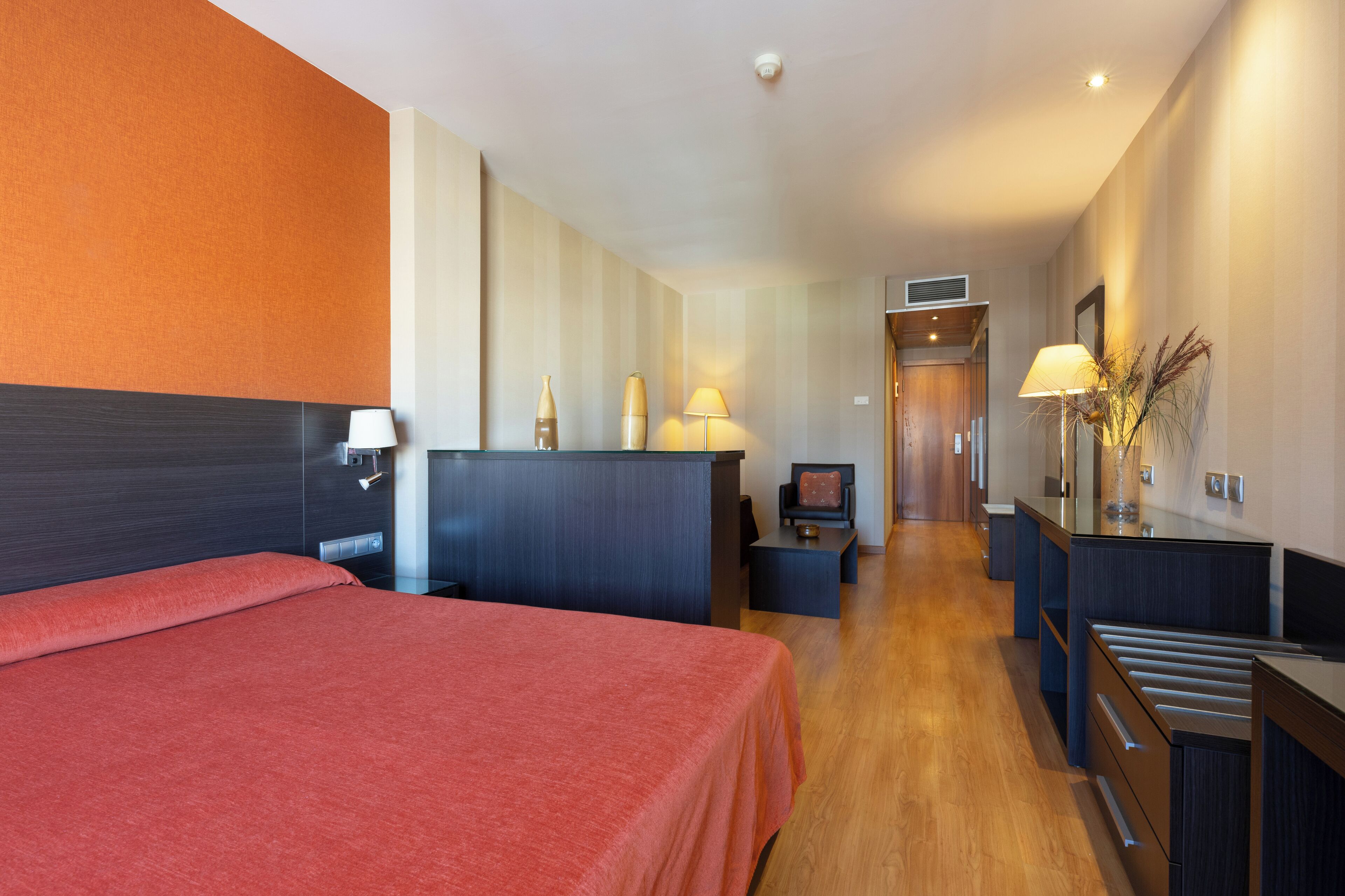A hotel room with a bed, a desk, a chair, a lamp, and a small table.