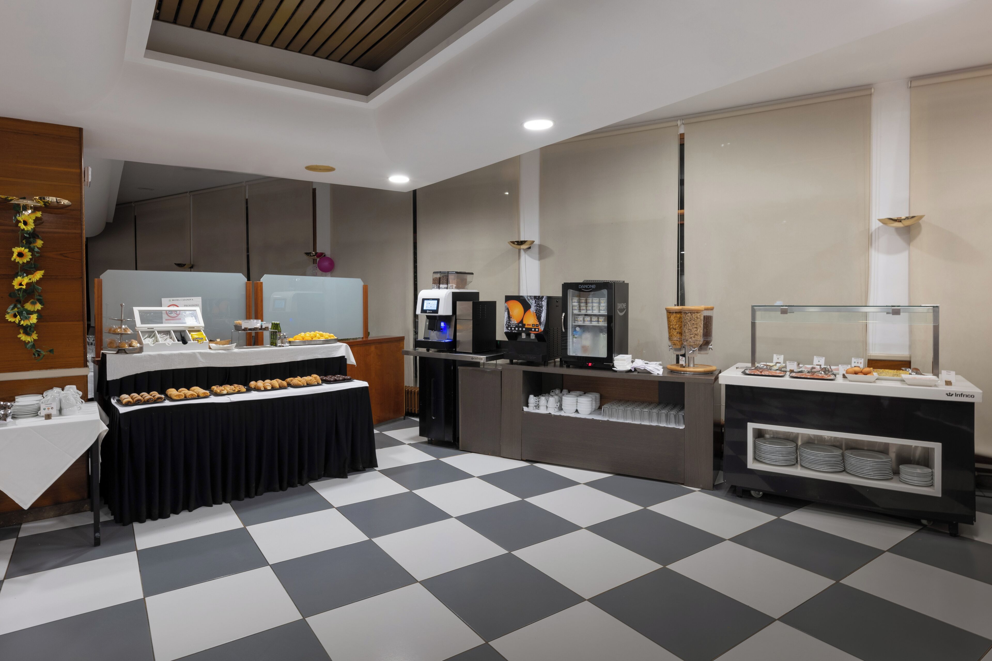 Een buffetopstelling met een geruite vloer, een lange tafel met etenswaren, een koffiehoek en een koelkast.