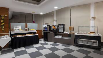 Een buffetopstelling met een geruite vloer, een lange tafel met etenswaren, een koffiehoek en een koelkast.