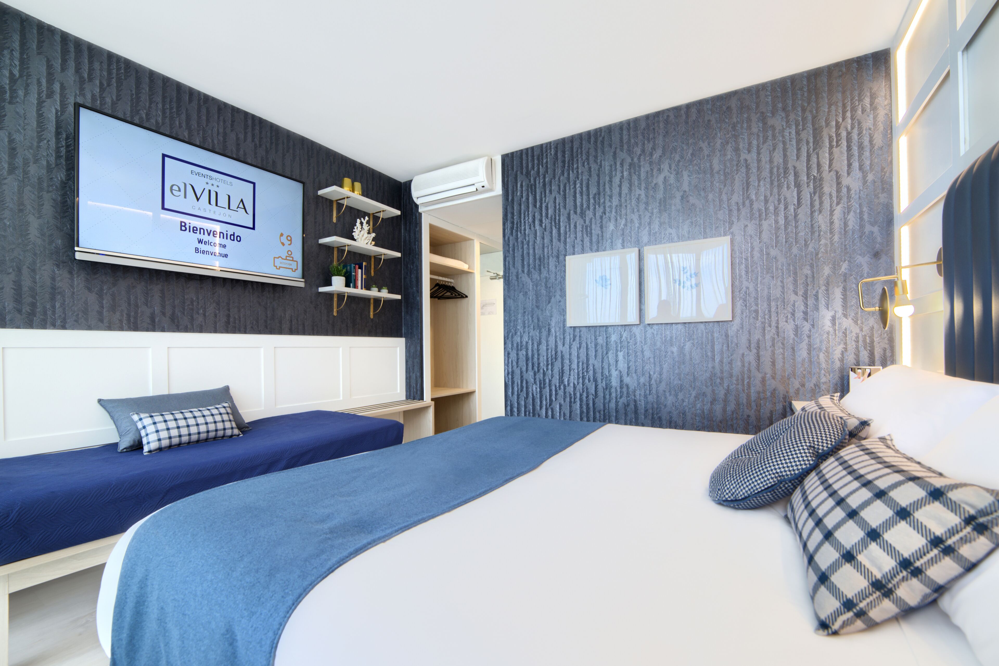 Habitación de hotel con una cama grande, dos camas más pequeñas, un televisor, un armario y una estantería con objetos decorativos.