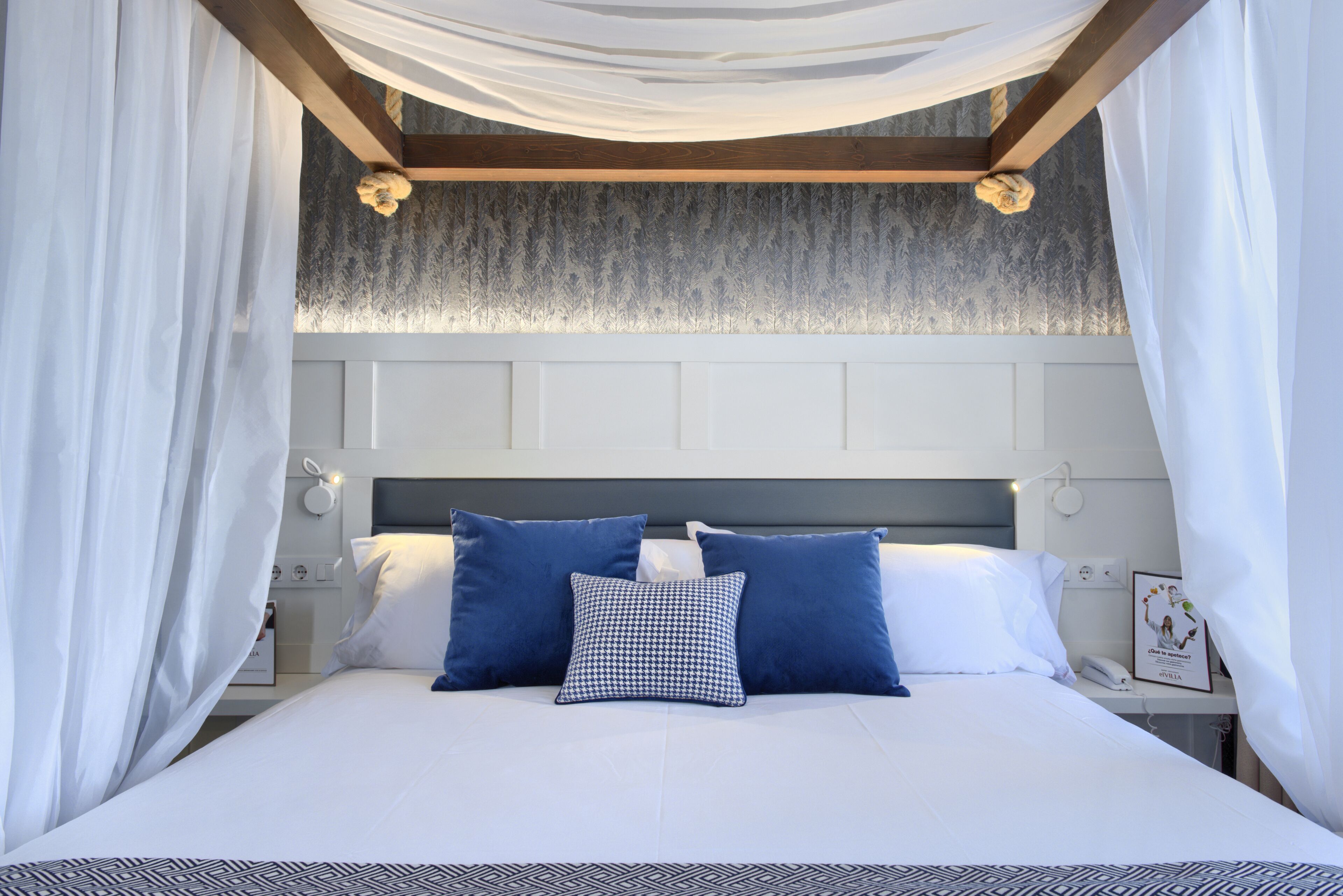 Una cama bien hecha con ropa de cama blanca y azul, un cabecero con papel tapiz estampado y una mesita de noche con una lámpara y una fotografía enmarcada.