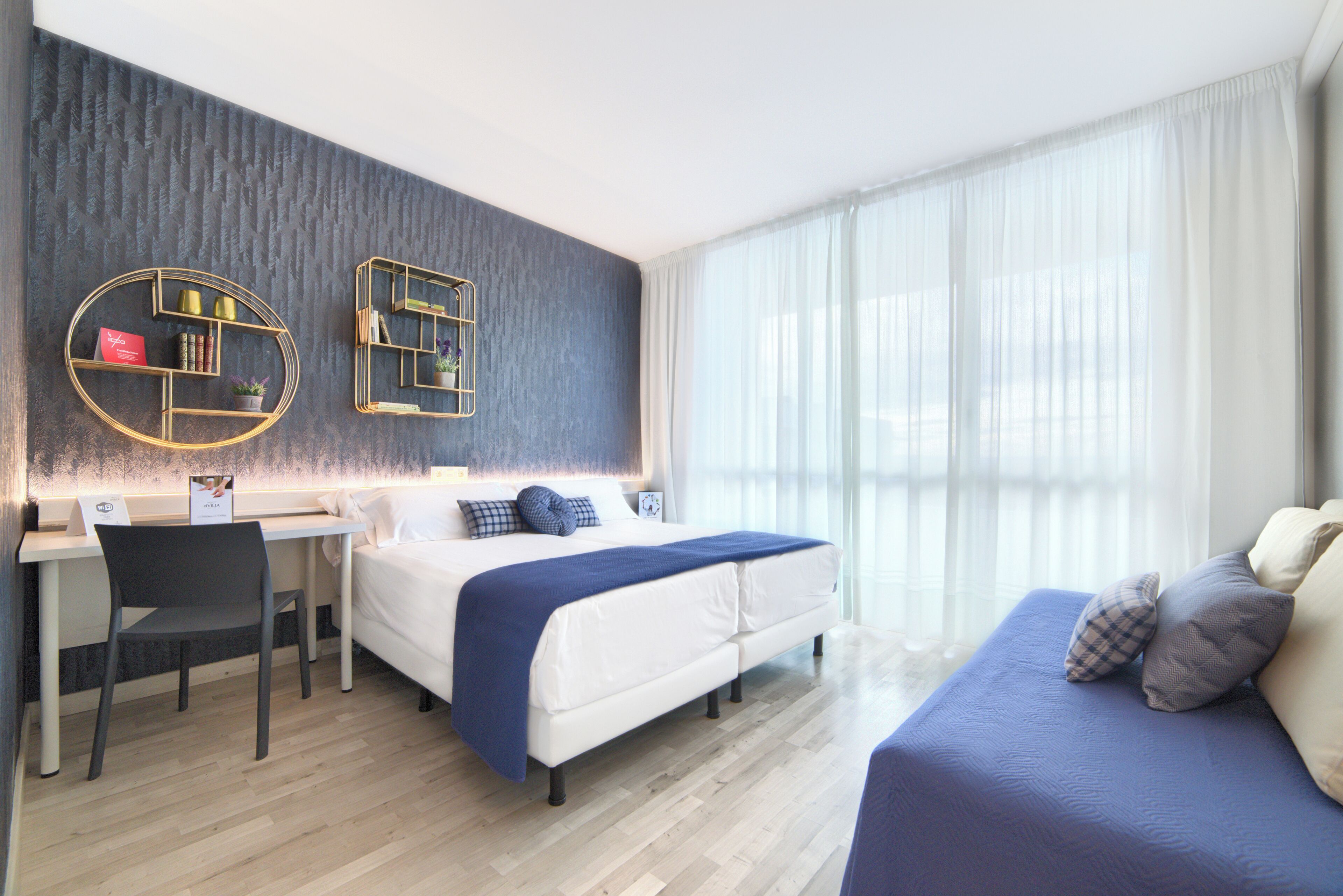 A modern hotel room with a bed, a desk, and a chair. There are two wall-mounted shelves with decorative items.