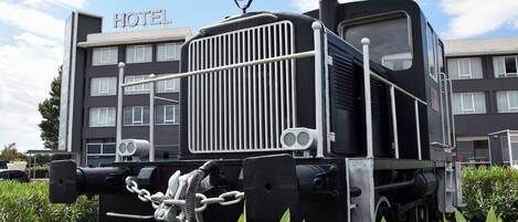 Una locomotora de vapor antigua frente a un edificio de hotel con múltiples ventanas y un letrero que dice 'HOTEL'.