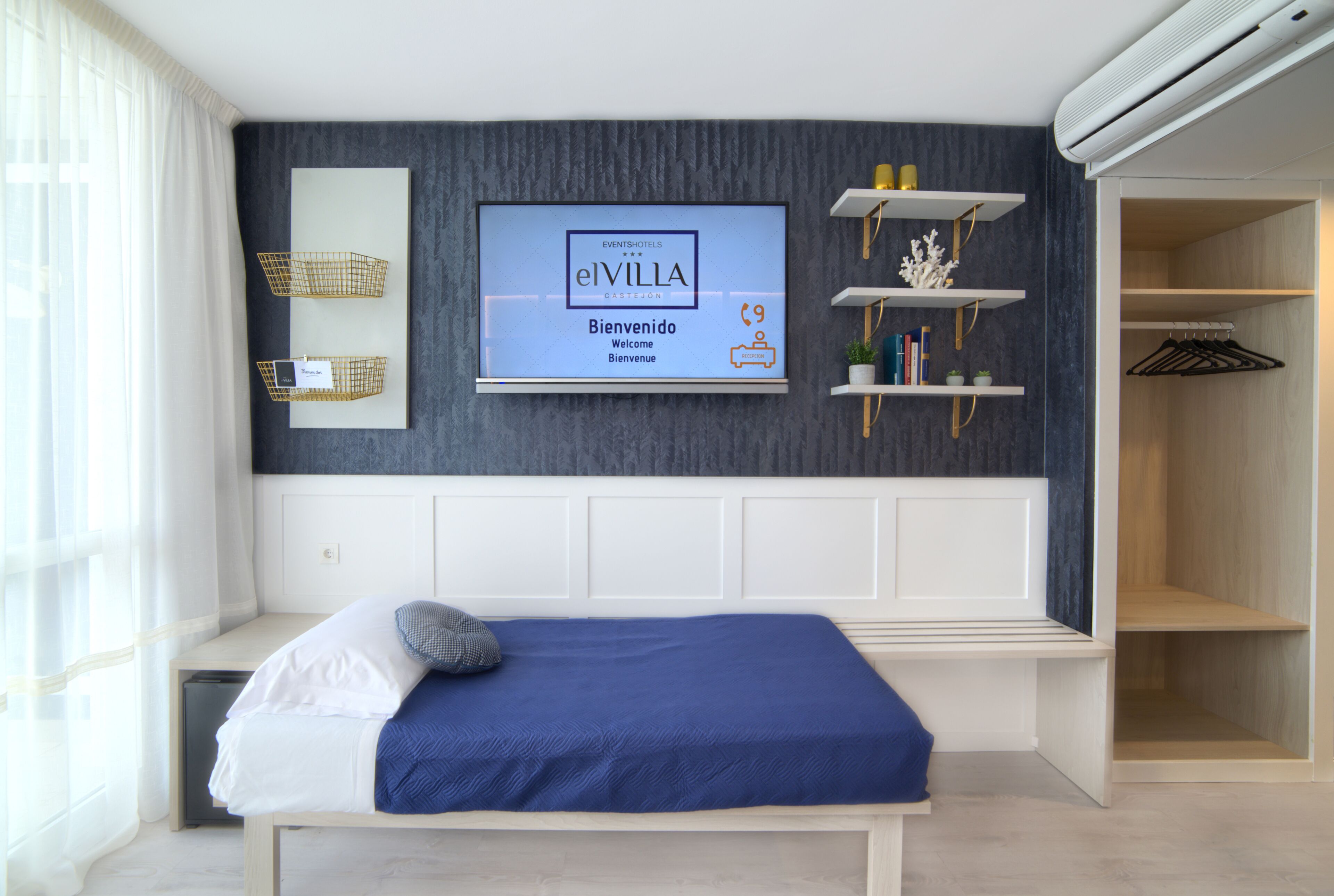 A modern hotel room with a bed, a mounted TV displaying 'elVilla' welcome message, built-in shelving, and a wardrobe.