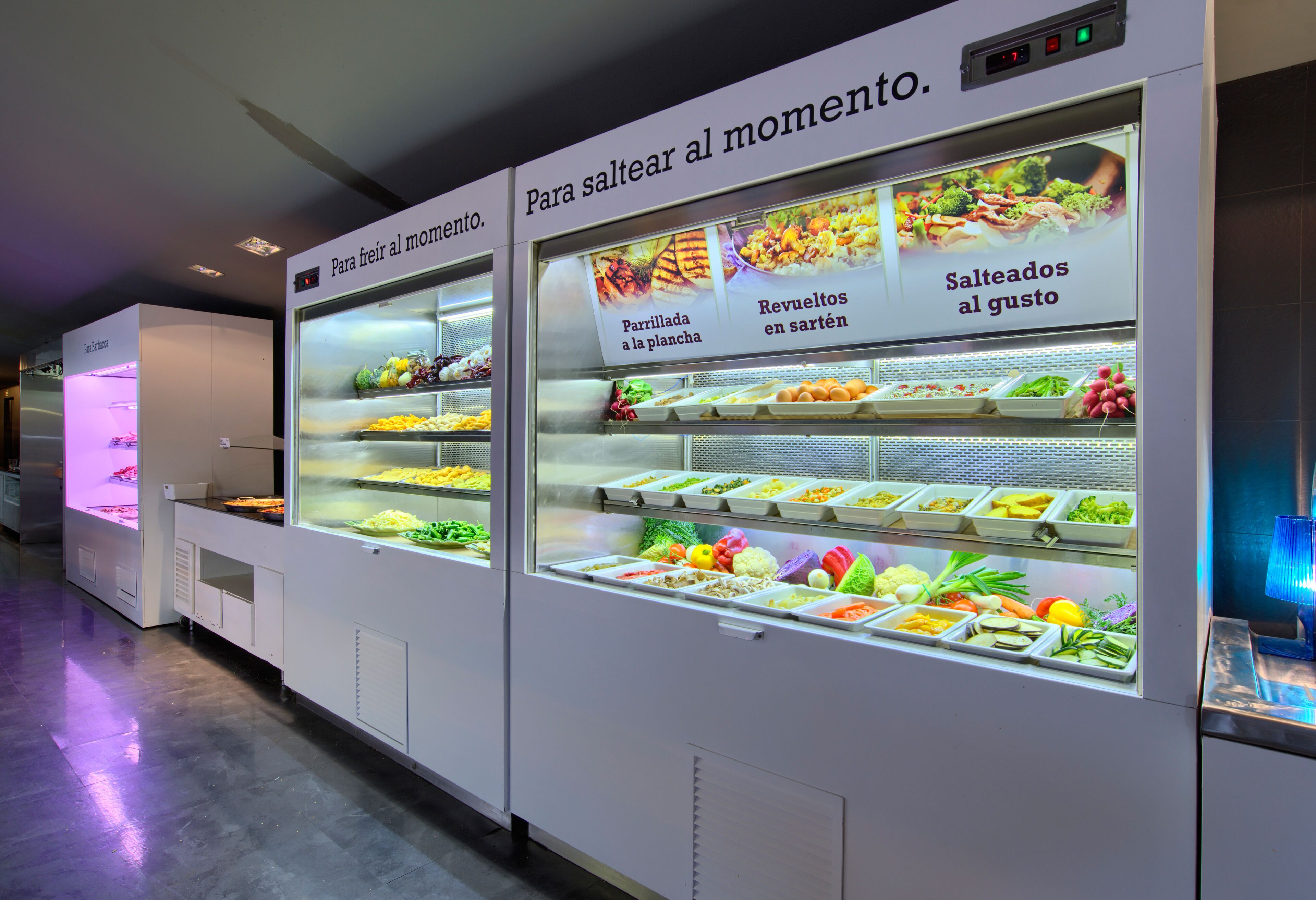 Esposizione di cibi con diversi frutti e verdure fresche, con etichette in spagnolo.