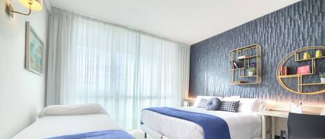 Una habitación de hotel moderna con dos camas, un escritorio y un gran ventanal con cortinas translúcidas.