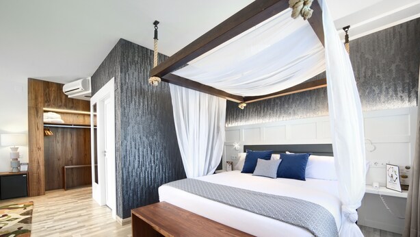 A modern bedroom with a canopy bed, wooden flooring, a bench, and a built-in wardrobe. - Hotel Luze El Villa (Castejón)