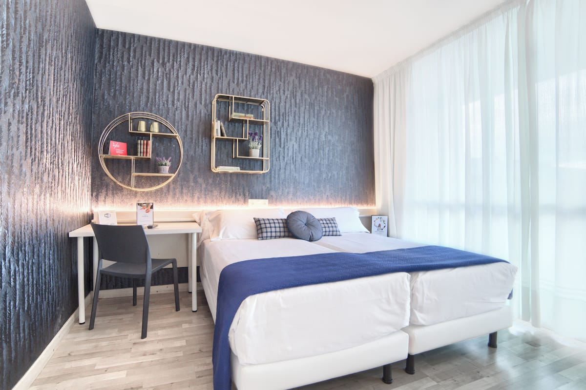 A modern hotel room with a large bed, a desk, and a chair. There are two wall-mounted shelves with decorative items.