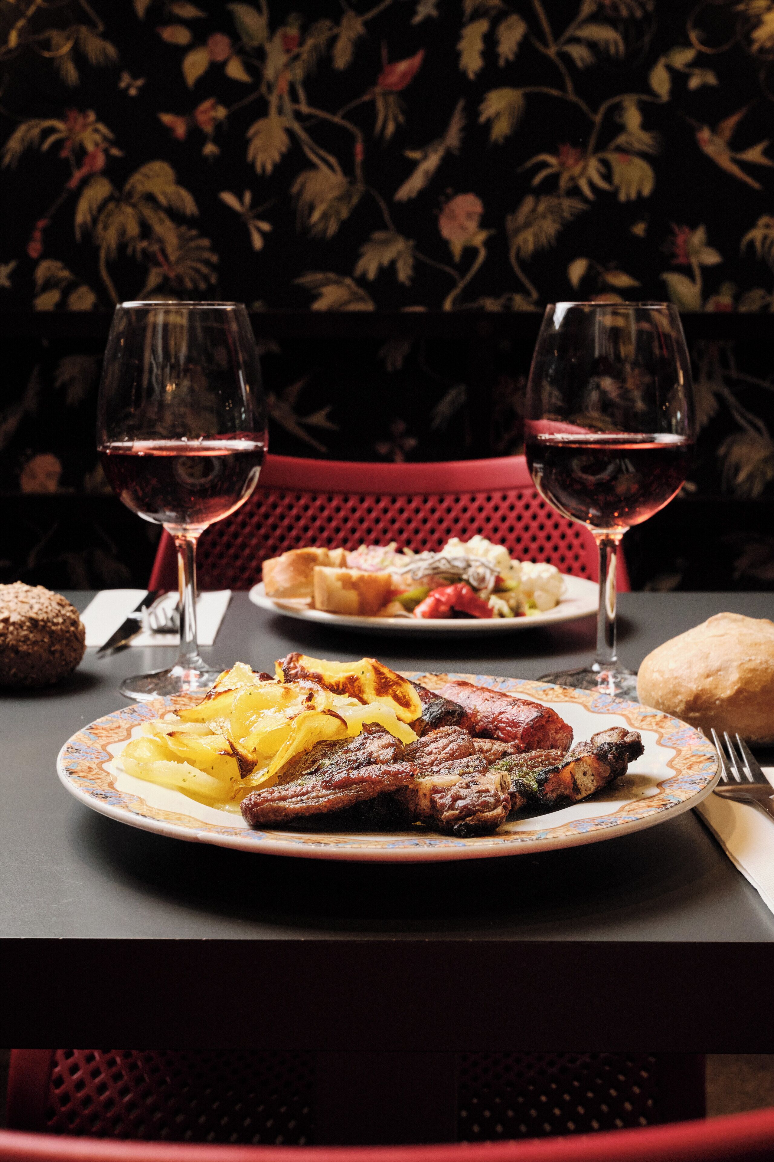 A dining table with plates of food, two wine glasses, and a red chair.