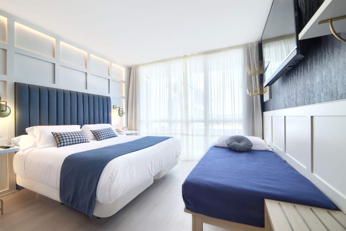 A modern hotel room with a large bed, a smaller bed, a desk, and a large window with curtains.