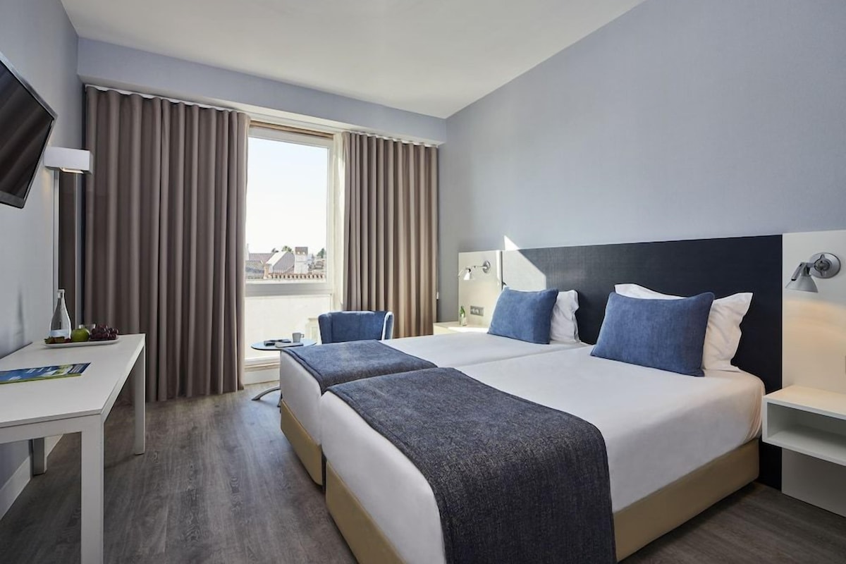 Quarto de hotel com duas camas, uma mesa, uma cadeira, uma televisão e uma janela com cortinas.