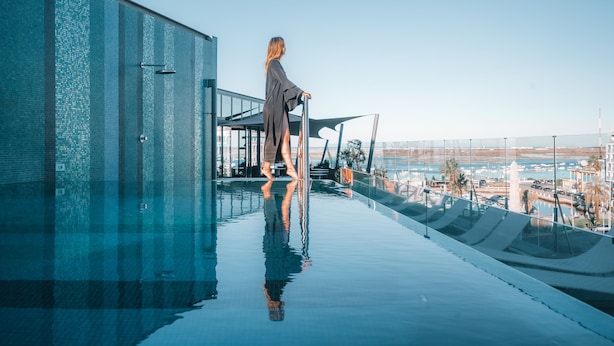 Une personne se tient près d’une piscine à débordement dont la surface réfléchit l’environnement, avec une vue sur un port de plaisance et des bâtiments en arrière-plan.