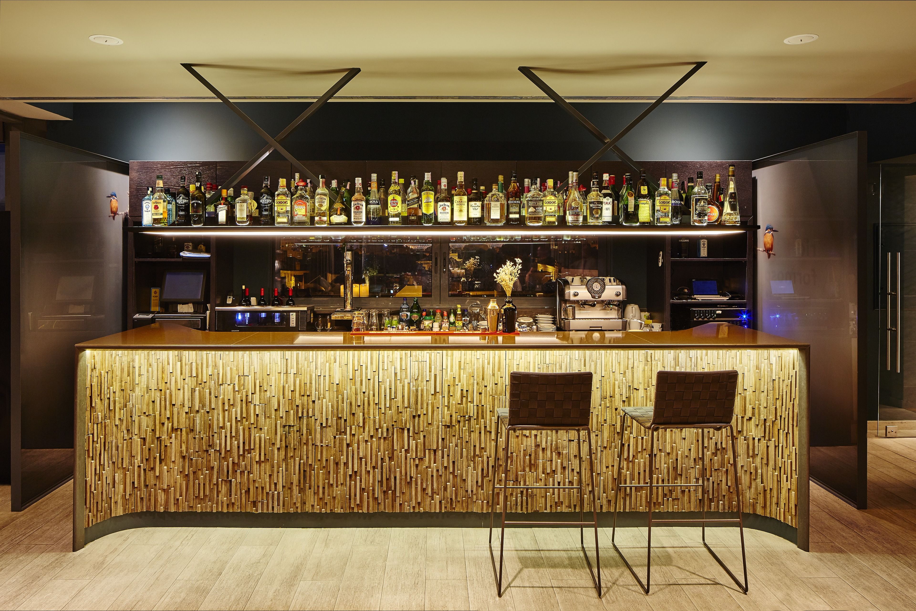 Un bar con diseño de madera, estantes con botellas de licores y dos taburetes.