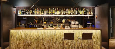 Un bar au décor en bois, avec des étagères remplies de bouteilles d’alcool et deux tabourets.
