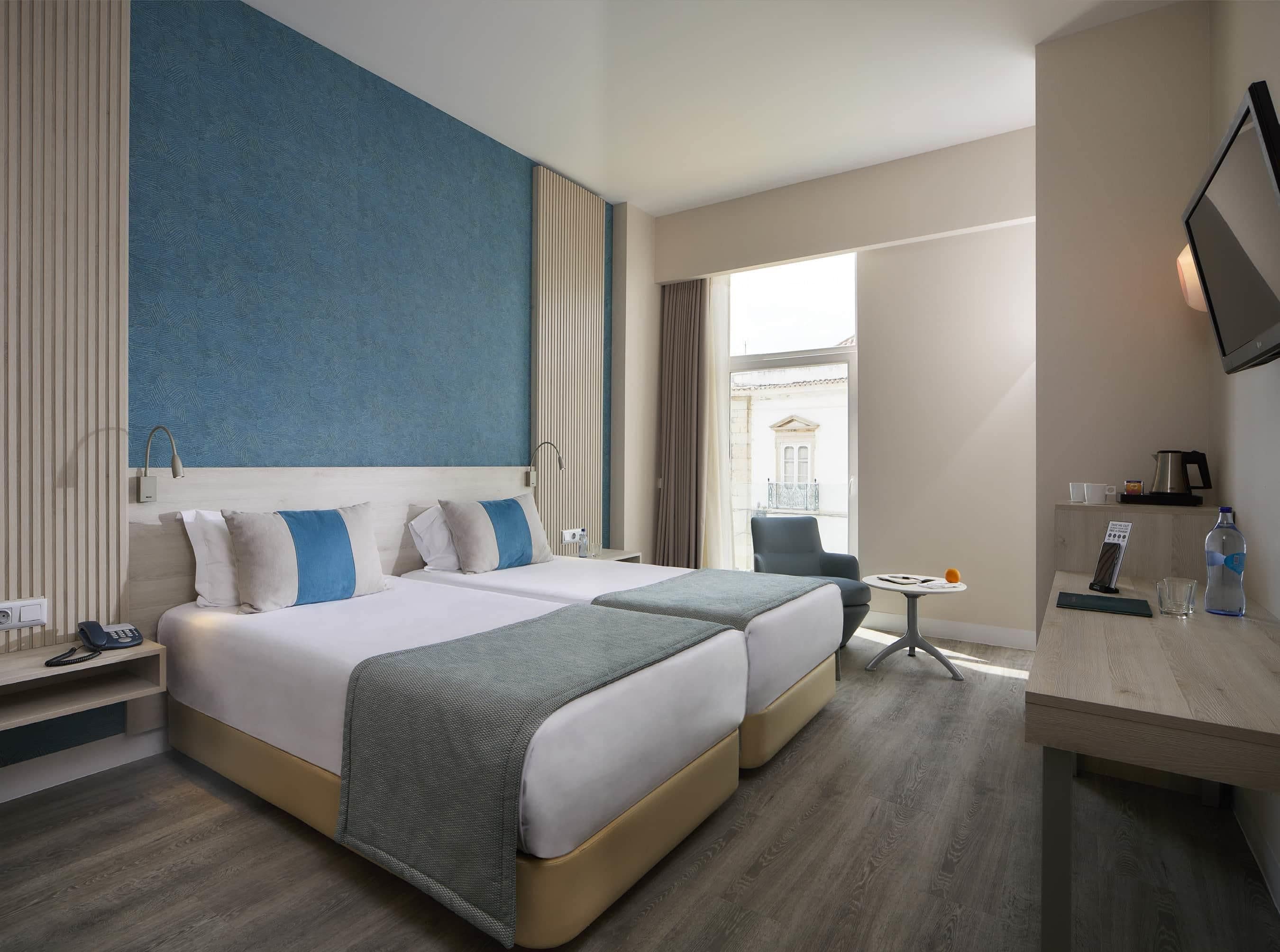 A hotel room with two beds, a desk, a chair, a window with a view, and a TV on the wall.