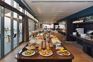 Disposición de un bufé con diversas opciones de comida y bebidas, una estación de café y vistas al exterior a través de amplios ventanales.