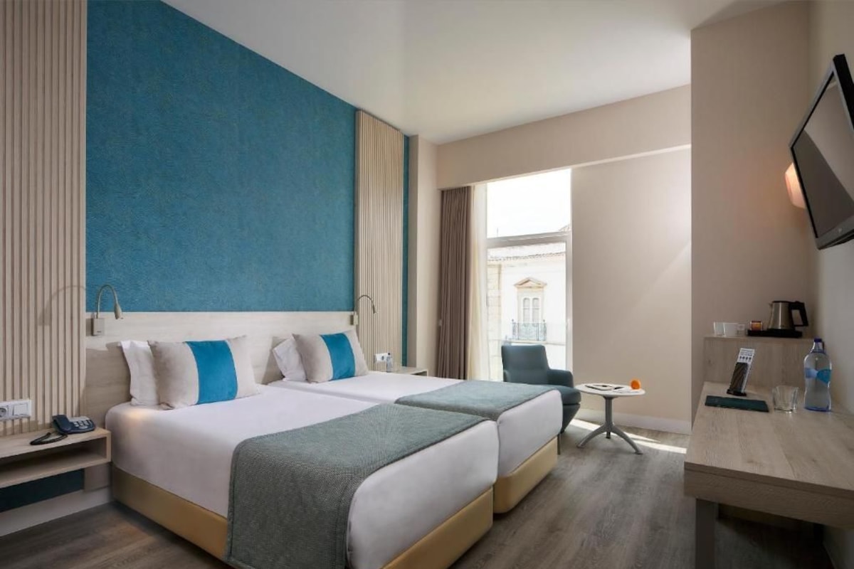 Quarto de hotel com duas camas, uma mesa pequena, uma cadeira e uma televisão.