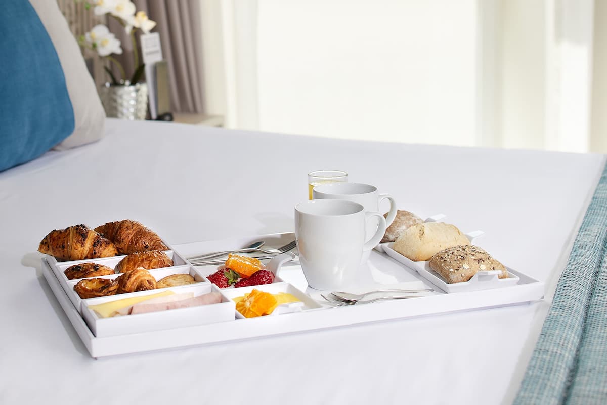 Una bandeja de desayuno ordenada con croissants, pasteles, fresas y rodajas de naranja sobre una mesa blanca en una habitación de hotel con vistas a edificios a través de una ventana.