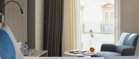 Une chambre d’hôtel avec un lit, une table sur laquelle se trouvent une bouteille, un verre et un livre. Il y a une chaise et une fenêtre donnant sur des immeubles.