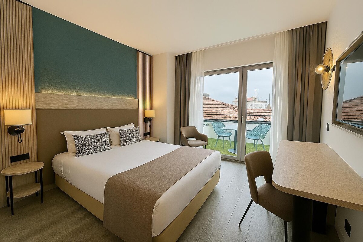 A hotel room with a large bed, a desk, two chairs, a small table, and a view of the cityscape through a large window.