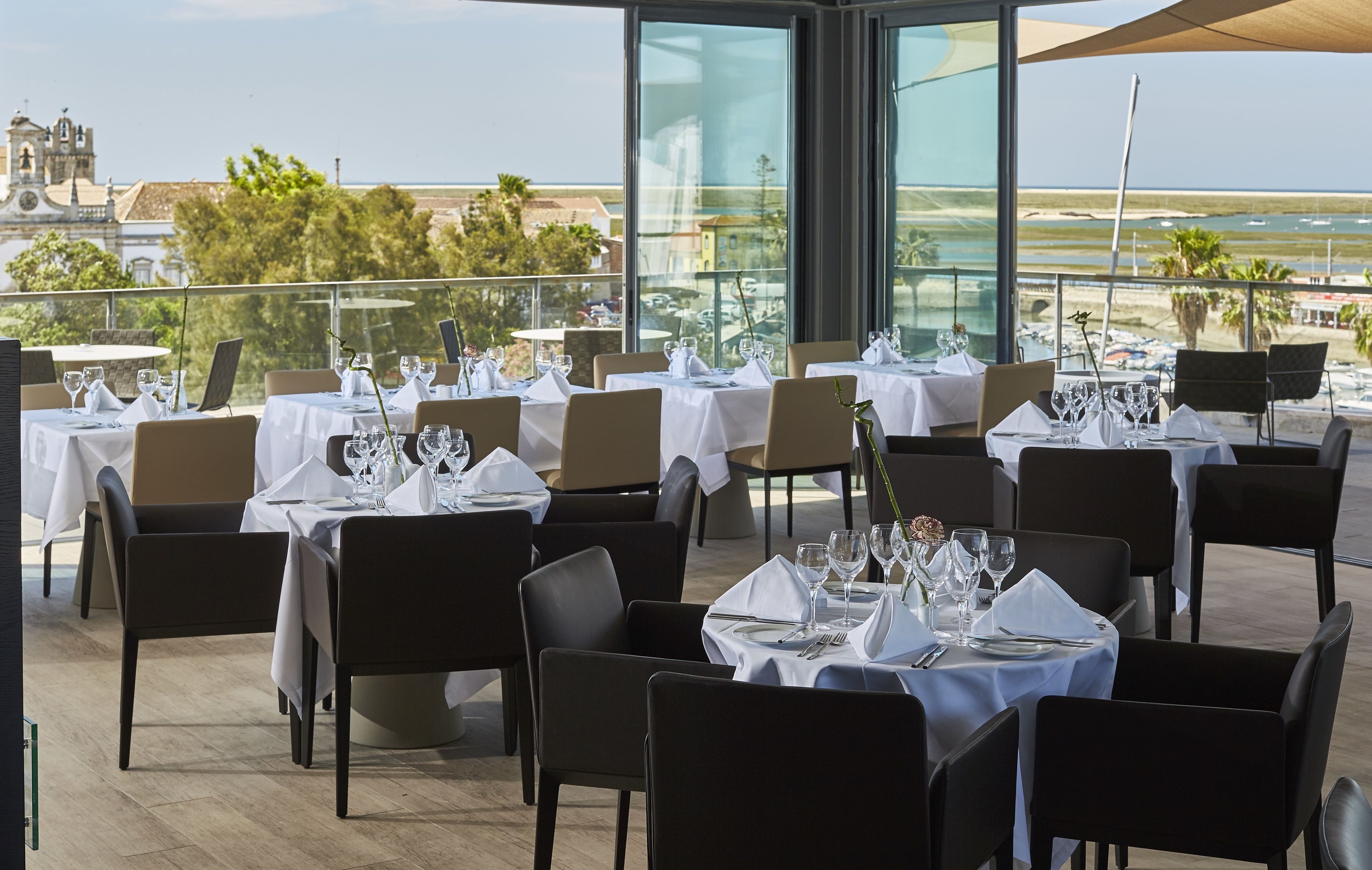A rooftop dining area with tables set for guests, overlooking a scenic view of the sea and buildings.