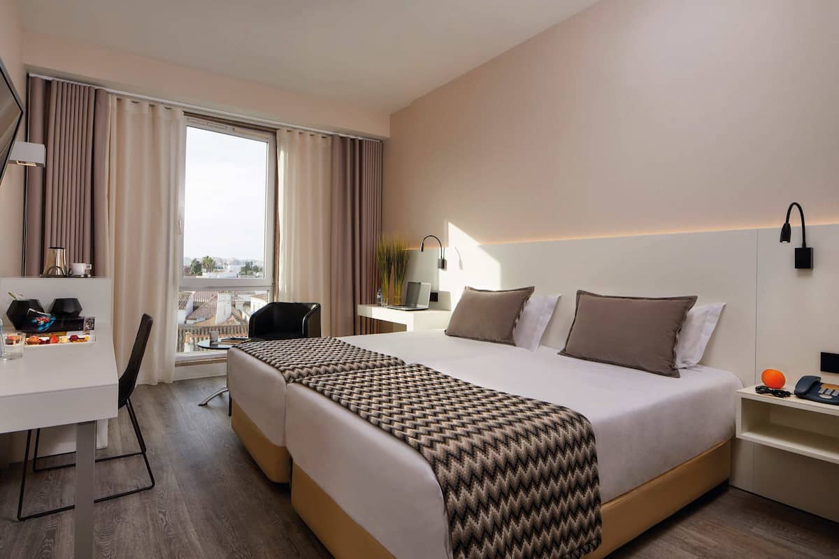 Una habitación de hotel con cama, escritorio, silla, mesita de noche y ventana con cortinas.