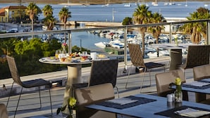 Une terrasse avec des tables et des chaises donnant sur un port de plaisance où sont amarrés des bateaux.