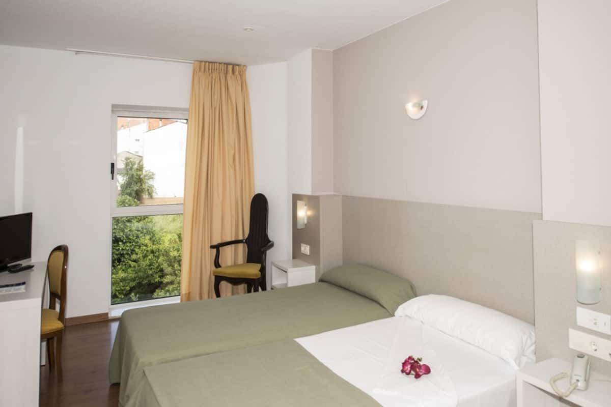 A hotel room with a bed, a desk with a computer, a chair, a window with curtains, and a view of greenery outside.