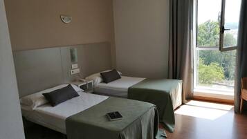 A hotel room with two single beds, a bedside table with a lamp, a power outlet, and a window with curtains.