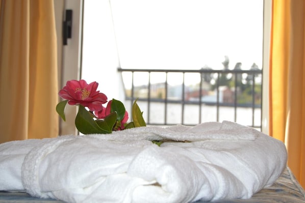 Una cama bien hecha con una toalla blanca, una flor rosa y vistas a un balcón con barandilla.