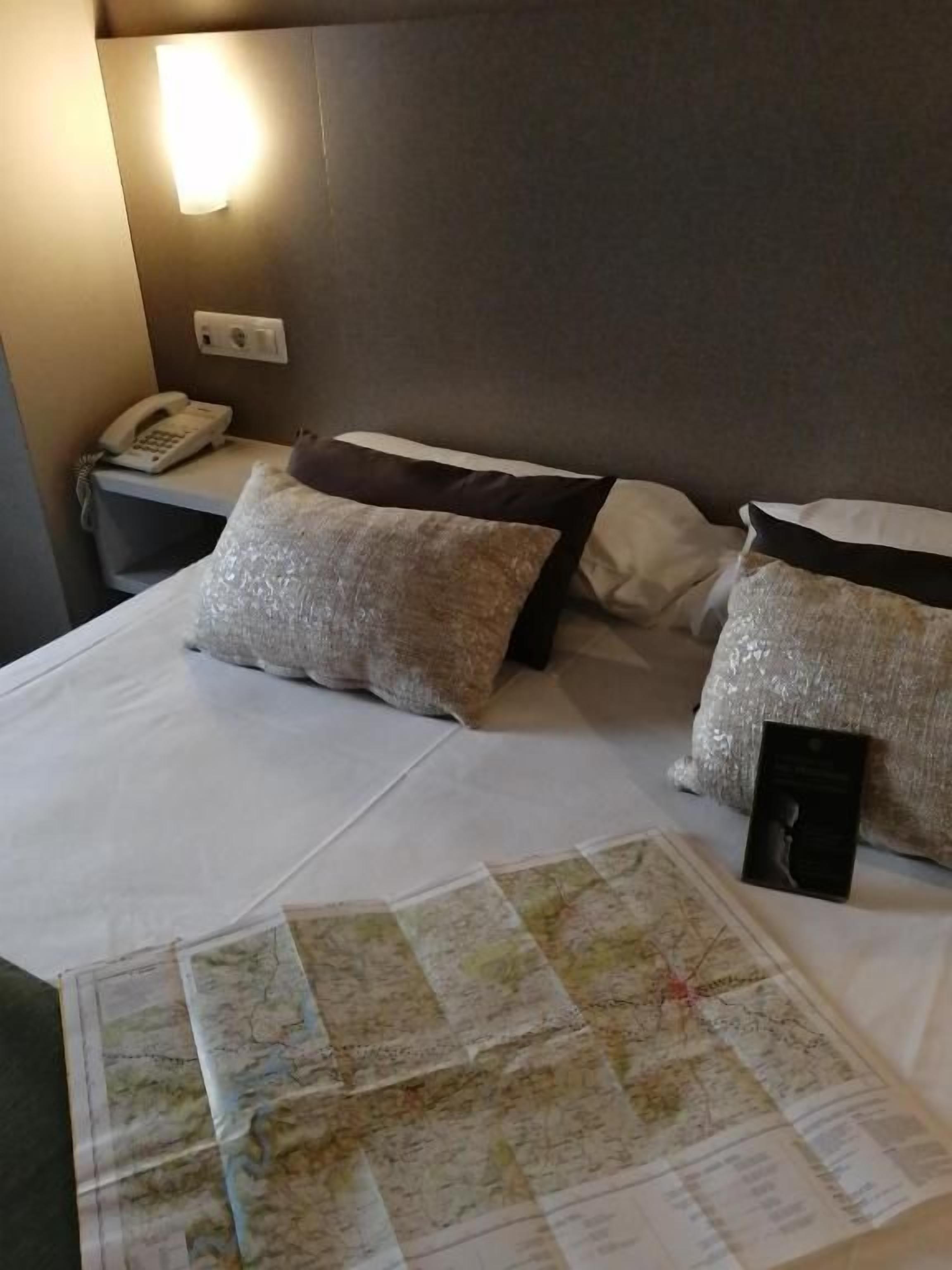 A hotel room with a bed, pillows, a map, a phone, and a wall-mounted light.