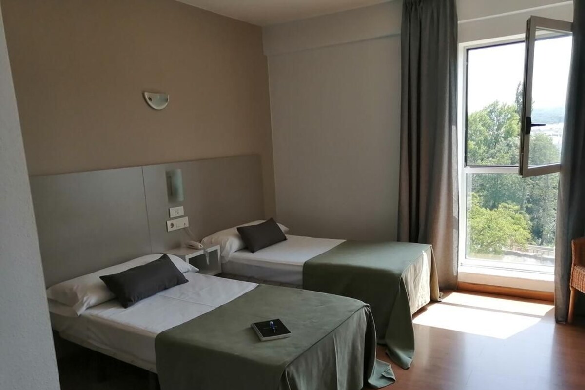 Habitación de hotel con dos camas individuales, un ventanal con vistas y una mesita con un libro.