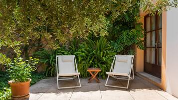 Dos sillones blancos con una pequeña mesa de madera entre ellos, ubicados en un patio con baldosas y un exuberante fondo verde de diversas plantas y un edificio con ventana.