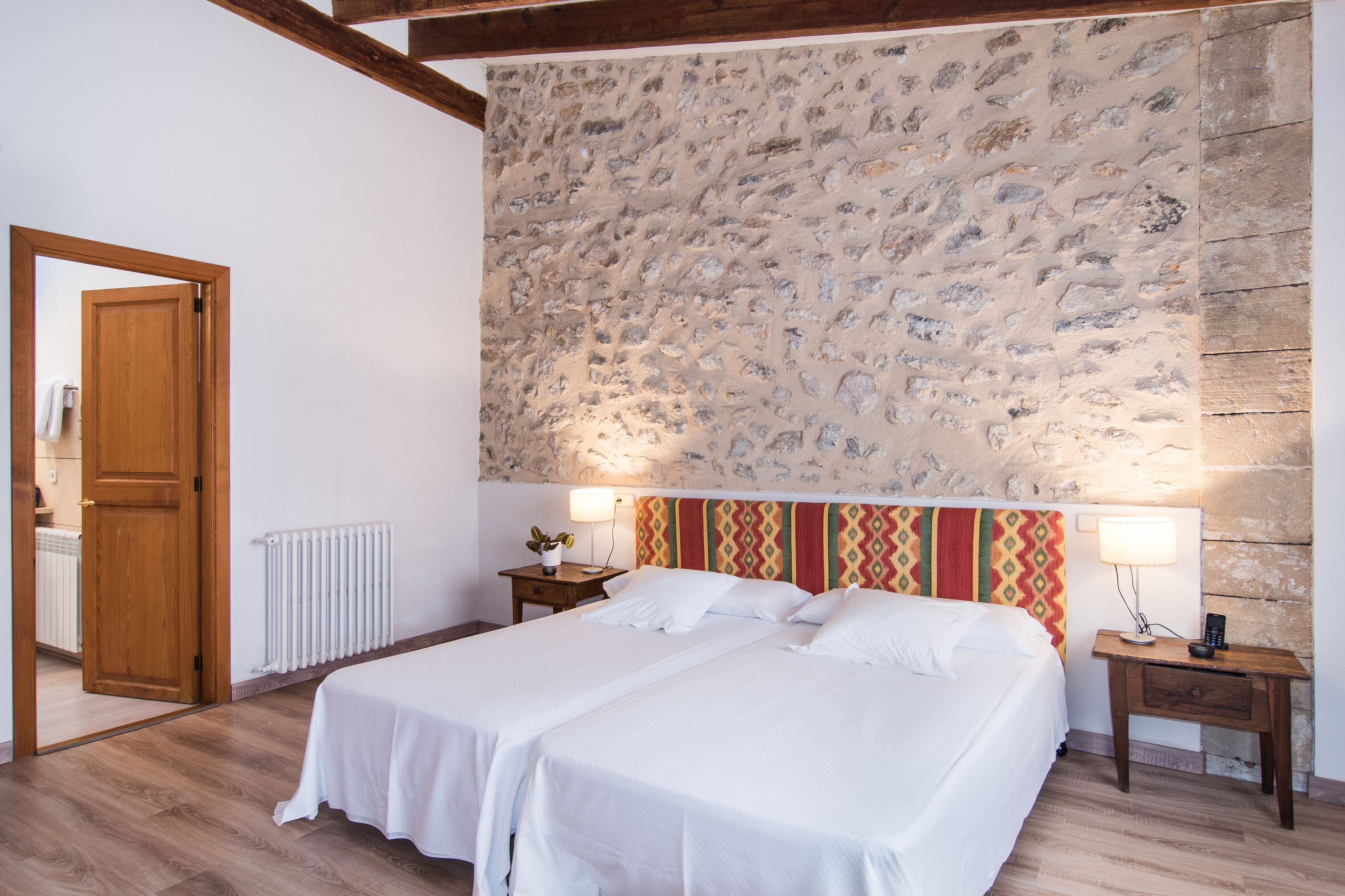 Una cama doble con sábanas blancas, una pared de piedra, una puerta de madera y una mesita de noche con lámpara.