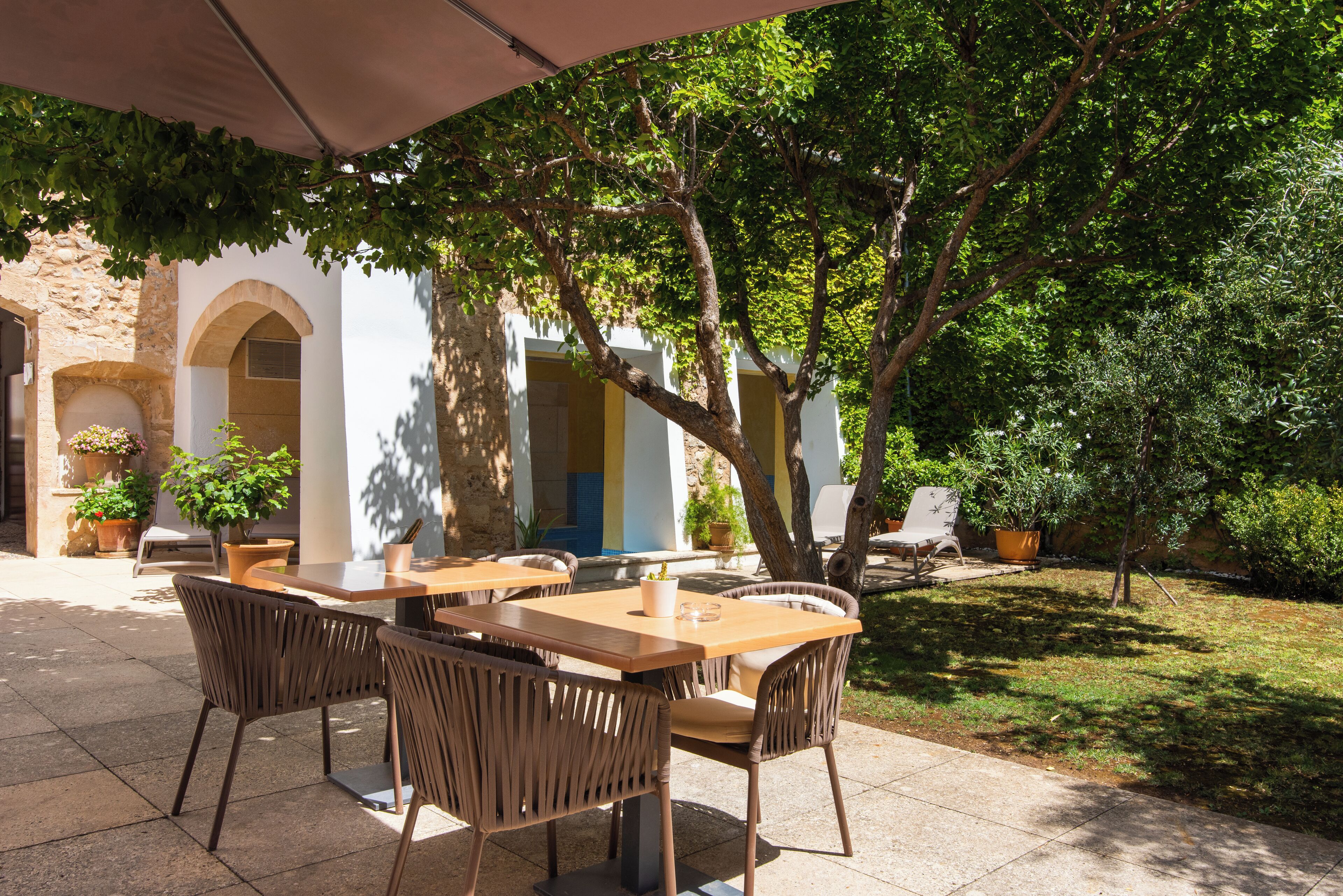 An outdoor patio with tables and chairs, surrounded by greenery and a stone building.