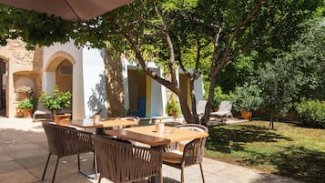 An outdoor patio with tables and chairs, surrounded by greenery and a stone building.