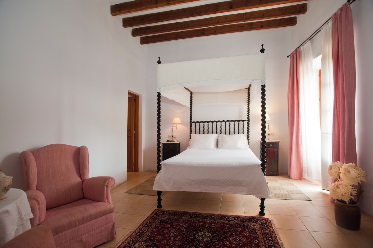 A bedroom with a four-poster bed, a wooden ceiling, a pink armchair, and a nightstand with a lamp.