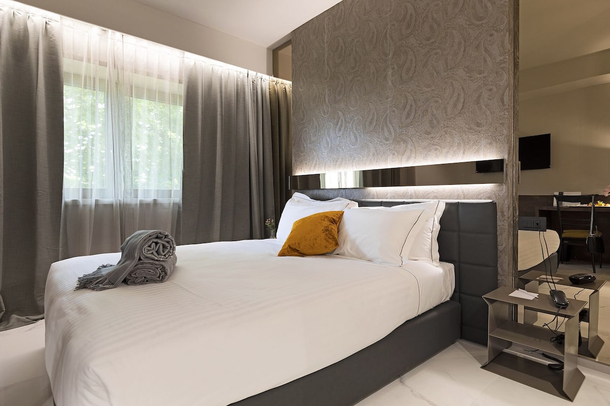 Habitación de hotel moderna con una cama grande, una mesita de noche con lámpara y vistas al exterior a través de cortinas translúcidas.